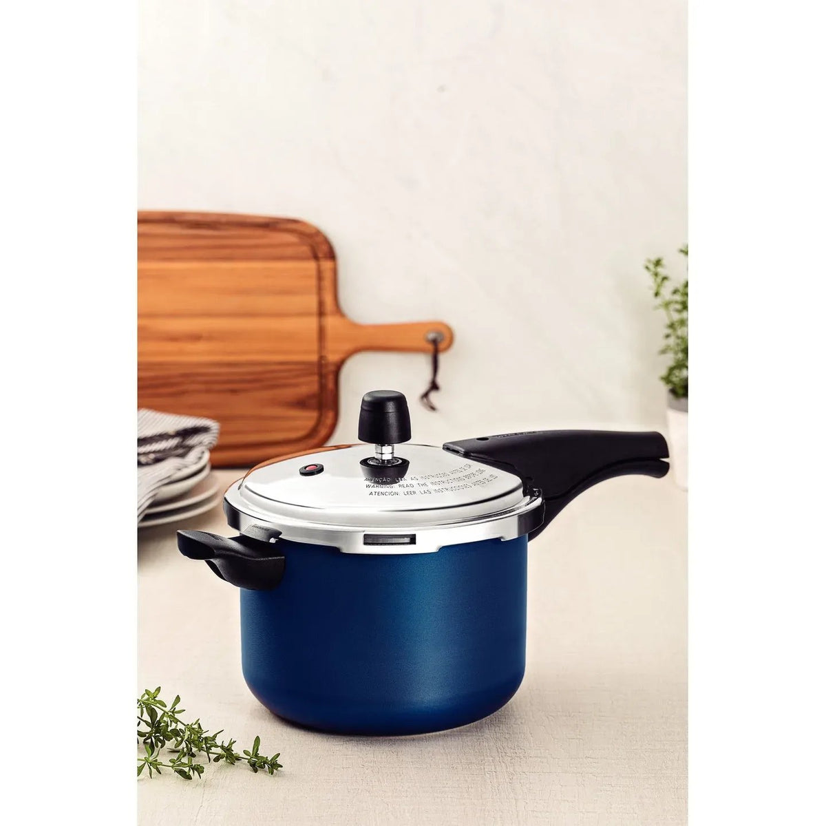 Olla de presión azul con tapa de acero inoxidable, válvula reguladora y mangos negros, sobre superficie clara con tabla