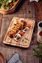 Tabla de madera con pinchos de carne, piña, tomate y cebolla, acompañada de salsa y romero sobre mesa rústica