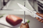 Cuchillo Tramontina de acero inoxidable con mango blanco siendo afilado con una chaira, sobre tabla de corte con carne