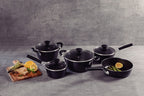 Juego de ollas y sartén negras con tapas de vidrio, acompañadas de tabla de madera con limones, queso y espárragos