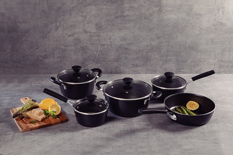 Juego de ollas y sartén negras con tapas de vidrio, acompañadas de tabla de madera con limones, queso y espárragos