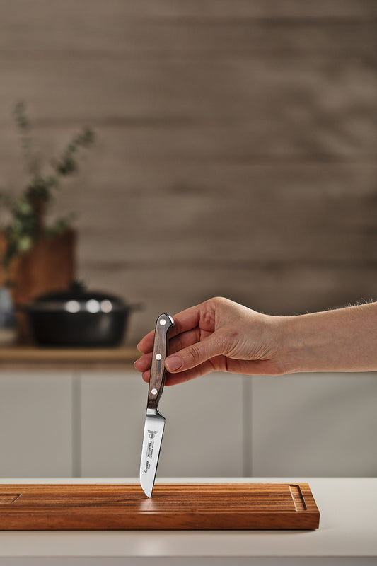 Mano sosteniendo cuchillo pequeño con mango de madera sobre tabla de cortar, con cocina al fondo